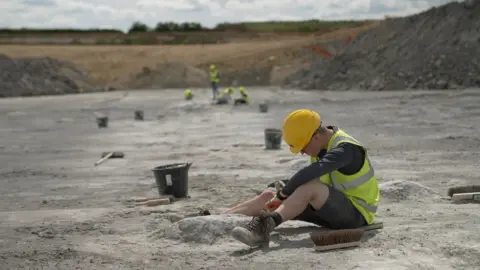 BBC/Kevin Church In a quarry of grey sand,  a man wearing a yellow hard hat, a yellow hi visibility waistcoat and shorts works on one of the footprints, which is a large crater in the ground. In front of him lies the brush of a broom without its stick. He seems to be digging with a small stick-like implement. A little away from him lies a bucket and what looks like a steel brush. Far in the distance and blurred out of focus, four more workers in hi visibility clothes do similar work, three sitting, one standing.