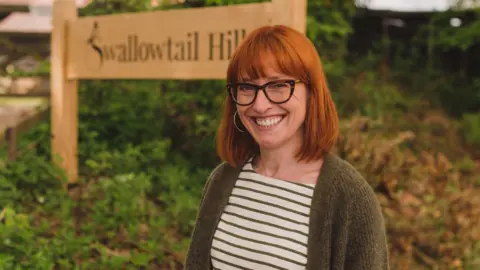 Sarah Broadbent Sarah Broadbent is wearing a white and brown striped top, a green cardigan and dark framed glasses. She is standing in front of a sign which says Swallowtail Hill