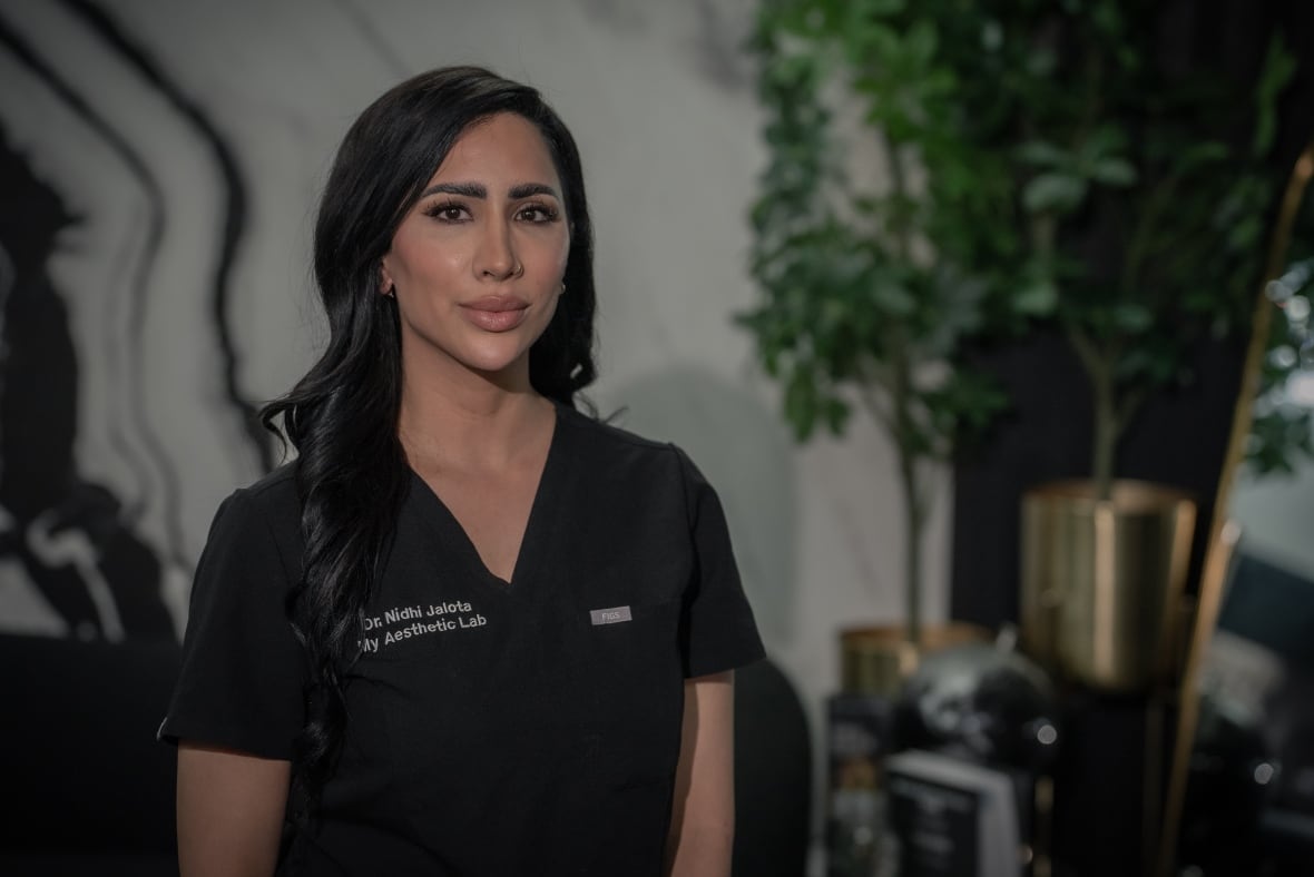 A woman with black hair wearing black scrubs stares straight into the camera