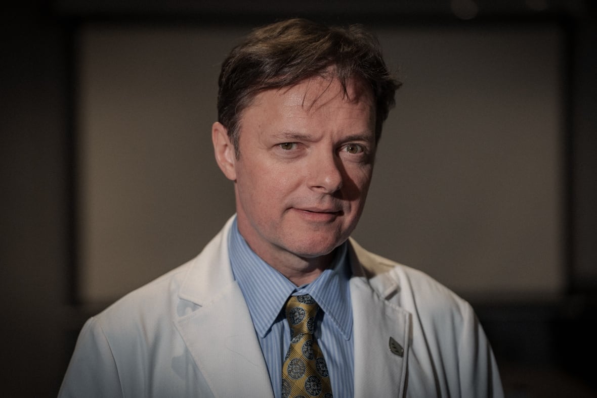 A doctor wearing a white coat with brown hair stares at the camera.