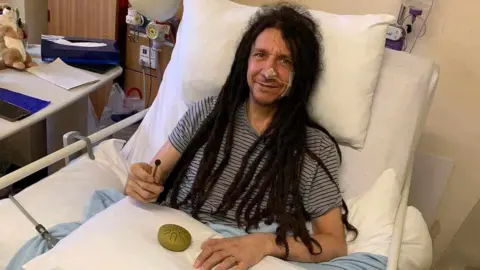 Tony Romaine Tony sitting in his hospital bed. He has a grey-T-shirt on, is propped up against the pillows in his bed, has medical and is smiling towards the camera. 