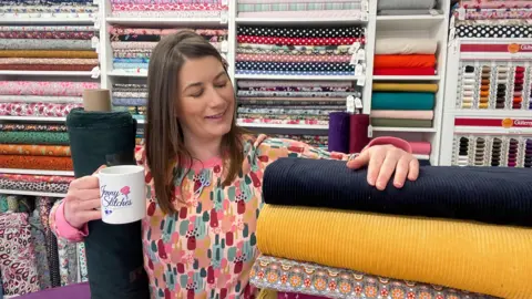 Jenny Fazackerly  A medium close up of Jenny Fazackerley, owner of Jenny Stitches in Barrow, stood her shop, checking fabric, and holding a mug of tea.