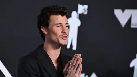 PA Media Shawn Mendes who has dark fairly short wavy hair and stubble. He is wearing a black jacket and looking towards his left with his hands up in prayer