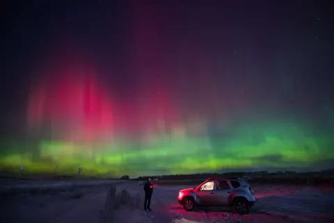 Alexander Manzyuk/Anadolu via Getty Images A man, holding a torch, stands next to a vehicle - with the interior light on -to observe the Northern Lights, appearing pink and green in the sky above the Siberian city of Krasnoyarsk, Russia on 2 January, 2025. 