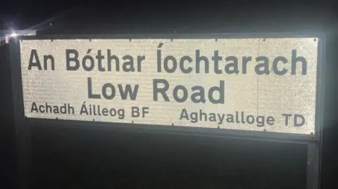 A white road sign with a black border which is reflecting the flash of the camera. The name of the road, where the incident happened, is written in Irish and English.