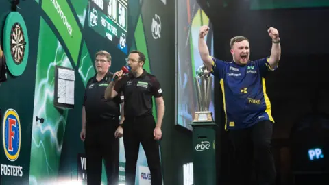 PDC Luke Littler celebrates on the right of the image with his arms in the air fist pumping towards the crowd. On the left, Huw Ware looks at the dartboard and holds a microphone to his mouth as he is about to announce the score. Another referee is stood to the left of him. The World Championship trophy sits in the centre of the image.