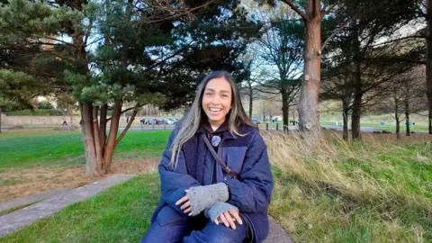 Devamsha Gunput A picture of Devamsha sitting in a park wearing a dark blue coat, surrounded by grass and trees.