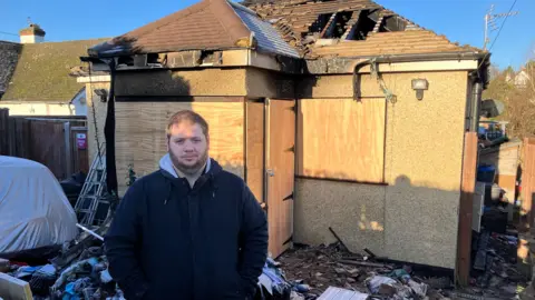 Toby in front of the remains of their former home. There are a few holes in the roof. There are various items and debris scattered around him.