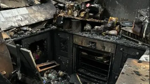 Toby The charred kitchen. The stove and utensils can be seen in the ashes.