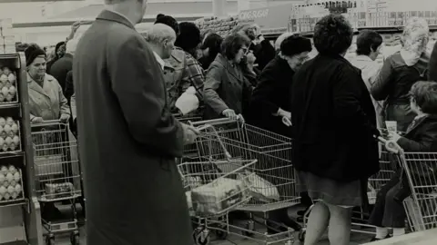 The Sainsbury Archive A black and white image of men and women in 1974 in a supermarket, pushing supermarket trolleys and looking at a just seen pile of sugar in front of them. Above it is a sign saying icing sugar