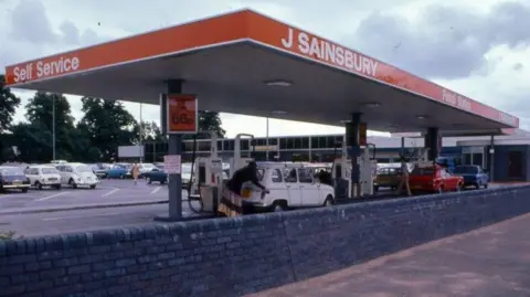 The Sainsbury Archive A petrol station behind a low wall in about 1975. It has a canopy roof branded in orange with self service, J Sainsbury and petrol station written in white lettering. Beneath the canopy a woman in a floor-length skirt can be seen putting petrol into a white car. Beyond the station is a carpark and a supermarket, with summer trees in leaf