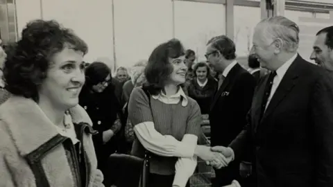 The Sainsbury Archive A black and white image showing women coming in through the entrance of a supermarket in 1975. One in the middle has shoulder-length hair and is wearing a short sleeved jumper over a white shirt and  is shaking hands with MP Jim Prior. He is wearing a dark suit and tie and has slicked-back grey hair