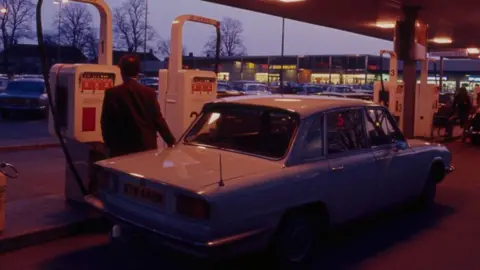 The Sainsbury Archive A back view of a man in a jacket standing behind a car and in front of a white petrol pump. He is filling the white car with his right hand. It is dusk and beyond the forecourt can be seen a Sainsbury's store and leafless trees framed against the sky