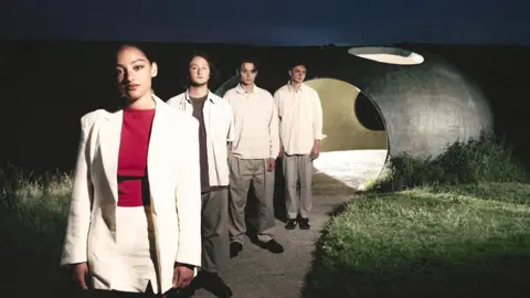 Tatiana Pozuelo Indie band English Teacher stand in a line while posing for a promotional photo near a hollow stone sculpture, lit from behind