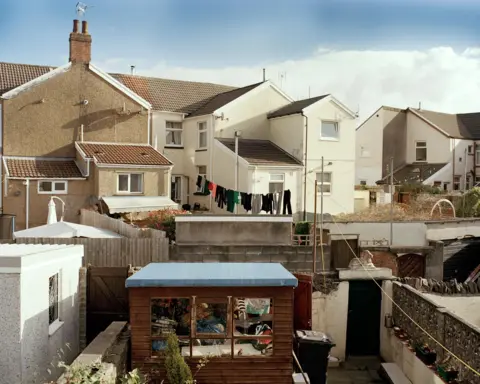 Megan Winstone A row of terraced houses photographed from behind with glimpses onto gardens, a shed and a washing line full of clothing 