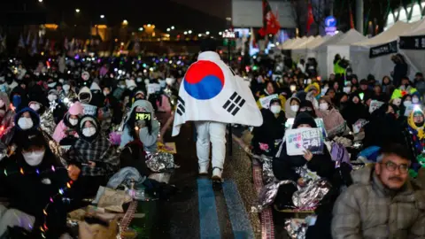 Getty Images A man wearing a South Korean flag as a cape walks through a sea of anti-Yoon protesters sitting on the ground.