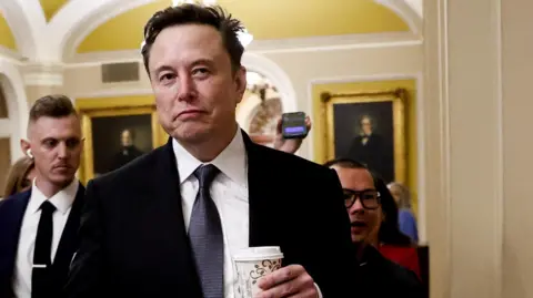 Reuters Elon Musk walking in Congress. The billionaire is holding a coffee cup as reporters follow behind him. 