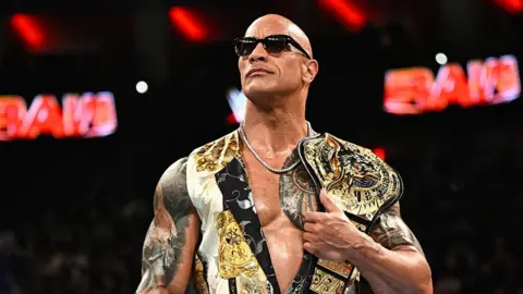 Getty Images The Rock standing in the middle of the ring, holding a golden title belt over his left shoulder, while wearing black sunglasses, a necklace chain and a white and gold top. Behind him is the "RAW" sign in red.