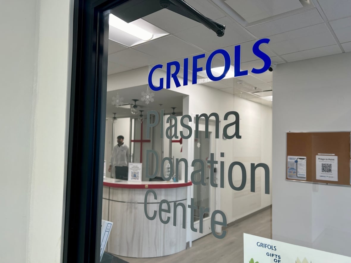 A glass door with the words Grifols Plasma Donation Centre, and a blurry man in the background walking toward a reception desk. 