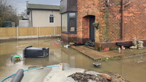 BBC/Simon Thake Floodwater surrounds a house with sandbags piled up at the door. A wheelie bin is tipped over on its side in the garden.