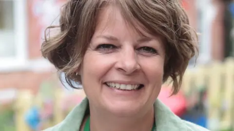 Caroline Gratrix Ruth Perry, who has short brown hair, looks straight at the camera in a portrait picture. She is smiling with a blurred background. 