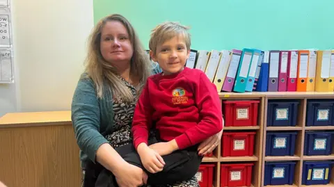 Hope Rhodes / BBC Penny and her son Thomas, sitting in a classroom. They both have blonde hair and are smiling.