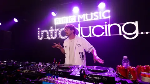 Barry Can't Swim behind DJ decks and other music equipment, with one hand spinning one of the decks and the other hand doing a dance move, and a large BBC Introducing logo behind him, at Radio 1's Big Weekend 2023 