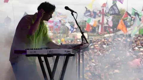 Side view of Barry Can't Swim on stage at a keyboard with a hand on his headphones, and a large crowd watching him and dry ice swirling around, on the Park Stage at Glastonbury 2024