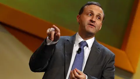 Getty Images A man with short dark hair wearing a dark grey suit, purple tie and white shirt giving a presentation at a political conference. His right hand is raised with his first finger pointing at something off-camera.