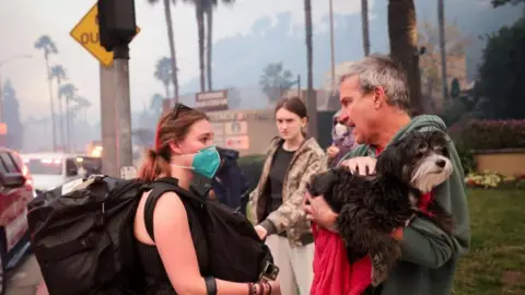 Getty Images A family, two young women and an older man, stand around waiting to be evacuated ahead of wildfires in LA. The man is holding a dog, one of the women has a facemask on. They have large bags.