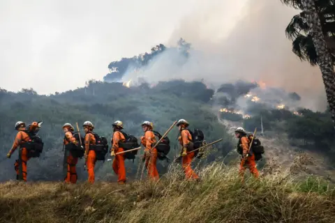 Caroline Brehman/EPA A group of firefighters walk through a clearing in woods with a burning hill in the background, in the Pacific Palisades.