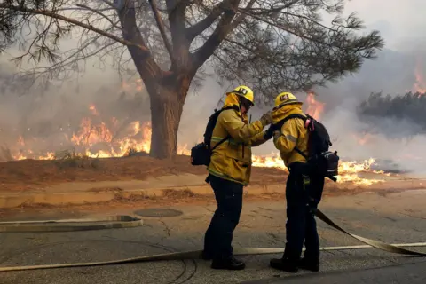 Caroline Brehman/EPA A firefighter helps their colleague wrap up in gear with burning forestry in the background in the Pacific Palisades.