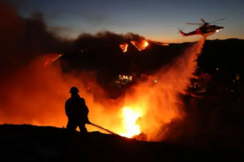 David Swanson/AFP A silhouetted firefighter fires a water cannon in front of huge flames in a valley with burning hills and a fire helicopter in the background in Pacific Palisades