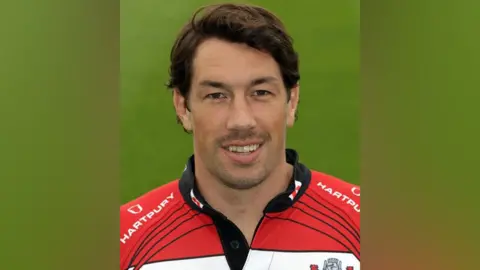 Getty Images A head and shoulders shot of Tom Voyce in a Gloucester rugby kit. It is a red and white hooped shirt with the logo of the company Jewson across the chest.