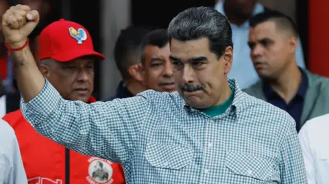 Reuters Nicolás Maduro wearing a checkered shirt hold up his right fist. Behind him, the interior minister, Diosdado Cabello, can be seen wearing a red jacket and a red baseball cap. 