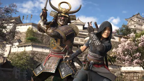 Ubisoft Screenshot shows a female ninja with a male character who's clad in elaborate samurai armour and horned helmet. They adopt battle poses as they stand in the gardens of a traditional Japanese castle. The building is visible in the background with cherry blossom trees blooming in front of its walls.