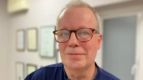 Dr Paul Charlson has grey hair, tortoiseshell-rimmed glasses and wears a navy blue shirt. He is pictured inside a room, painted white, with frames up on the wall behind him.