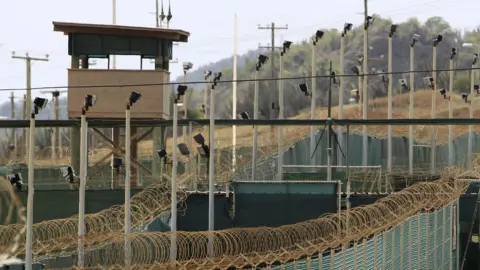 Reuters Aerial photo the prison at Guantanamo Bay shows high fences topped with barbed wire, several cameras on poles and a watch tower. There are trees and brush in the background as well as another fence