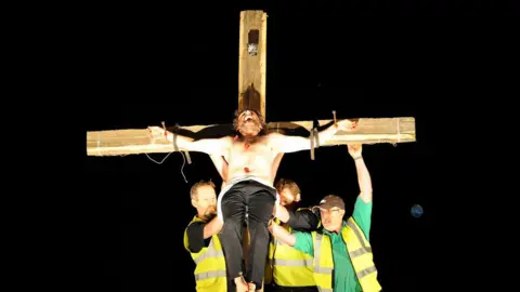 BEN STANSALL/AFP via Getty Images Michael Sheen topless tied to a crucifix, with three men in high vis jackets propping him up