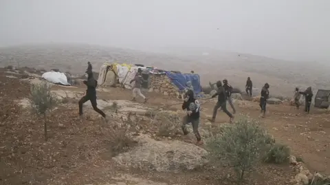 B’Tselem About a dozen people dressed in black with faces covered by hoods and scarves run in the same direction across dry ground, with a small stone building in the background, Khirbet Susiya in the South Hebron Hills, 21 December 2024