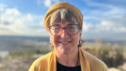 Standing outside, Sondra stares into the camera, wearing red rimmed glasses, a yellow scarf and yellow cardigan. In the background is out of focus greenery