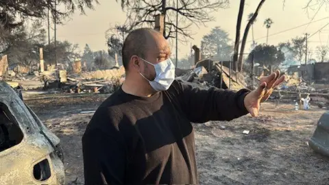 Larry Villescas stands in front of the charred remains of his neighbourhood. 