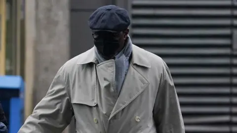 PA Media Charles Ehikioya wearing a beige trench coat, grey scarf, glasses, a black mask covering his face and a grey hat.