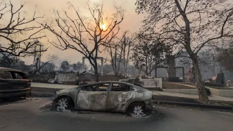 Lucy Sherriff / BBC A burned out car surrounded by burnt buildings as the sun shines through smokey clouds