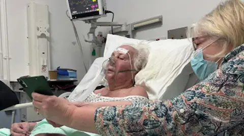 Family handout An elderly lady, Mary Lamerton is lying on a trolly in A&E. There is a lady sat next to her in a mask who is holding a phone. 