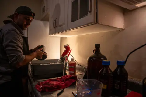 Fritz Pinnow Fabriccio Díaz, wearing a back-to-frong baseball cap, is moving the container with the developing film around for the chemicals to mix evenly with the film roll.