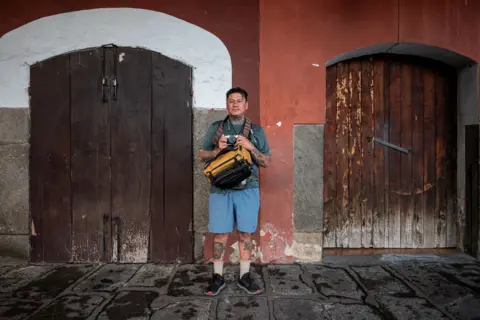 Fritz Pinnow Steven López, wearing shorts and carrying a camera bag, stands in front a building on his way to take photos on his travel across Central America. In his hands, he holds his trusty analogue camera