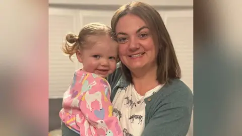 Gemma Whatling A young girl with blond hair and a pink jumper with horses on is hugged by her mum, who has short brown hair and is wearing a grey/blue cardigan and white t shirt