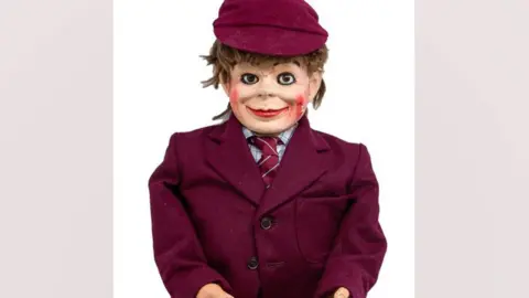 Hansons Auctioneers A head and torso shot of Eric Morecambe's ventriloquist dummy Charlie. It is wearing a maroon jacket over a checked shirt and a maroon tie. It has a maroon cap on its head, from which light brown hair is sticking out. It has a broad smile