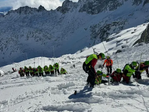 Soccorso Alpino e Speleologico Trentino About a dozen rescuers in high viz jackets using equipment search through snow in mountains